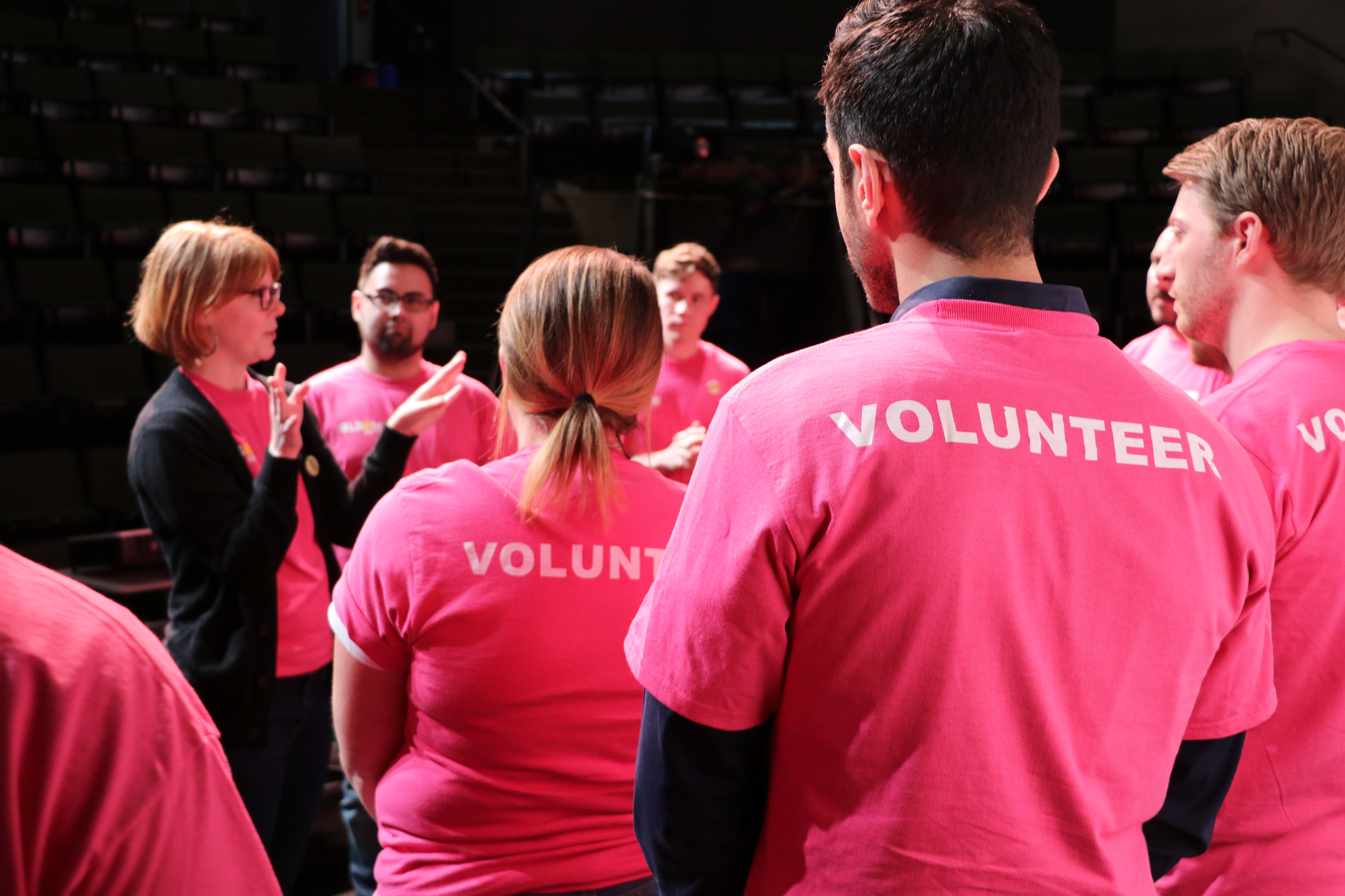 At a GLSEN Greater Cincinatti event, several adult volunteers in pink shirts circle around a speaking volunteer in a black cardigan. The back of the pink shirts reads: VOLUNTEER. 