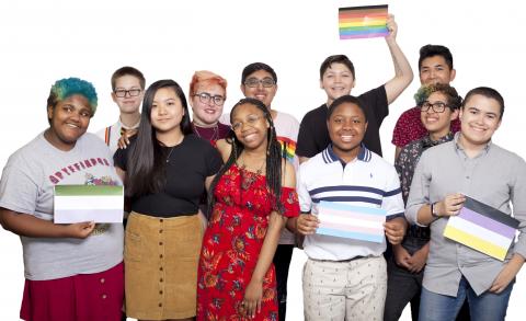 The 2018 National Student Council members stand in a group smiling at the camera in front of a white background. Several hold up print-outs of various pride flags. From left to right, they are: genderqueer, rainbow, trans, and non-binary. 
